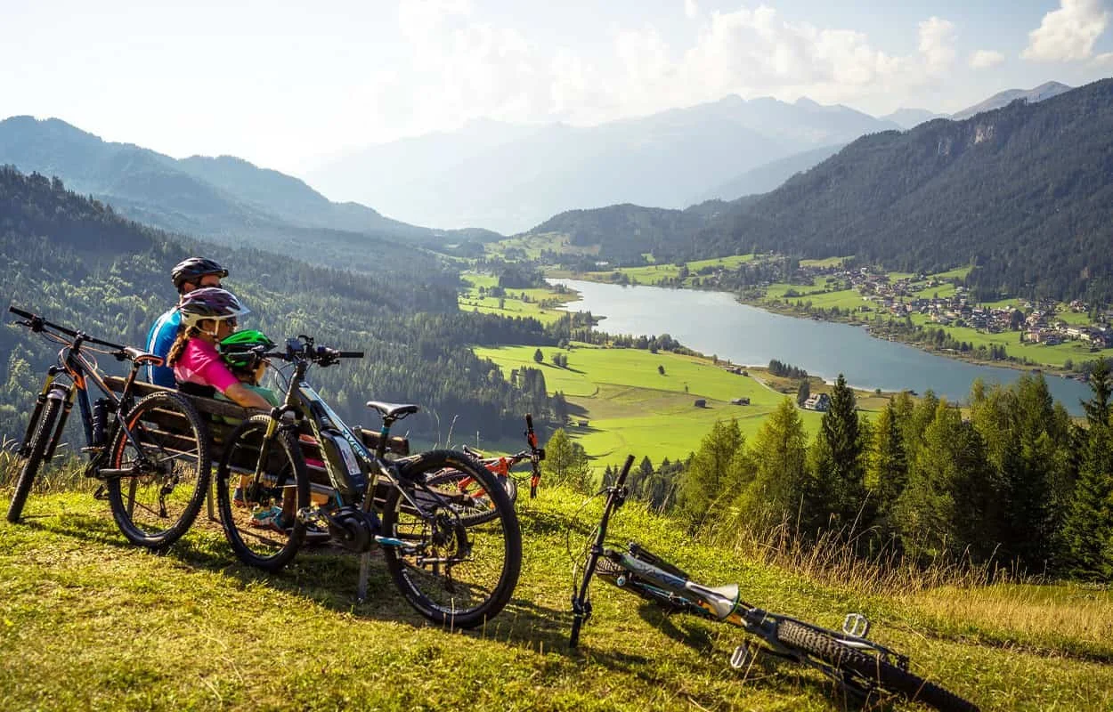 Two children with helmets sitting on mountain bikes overlooking a green valley with a lake.